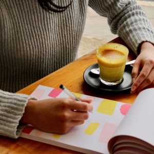 A black woman is writing on a pad that has colored blocks on it. She has a coffee on the table.