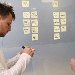 a Caucasian make standing at a board with sticky notes on it as he writes on a sticky note