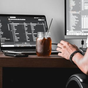 A dark brown desk with a black laptop that has a black background and white words on it. Outside of the laptop is a glass drink.
