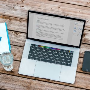 A outside table with a laptop opened to a document, a glass of water and a folder with a document on top is on it.