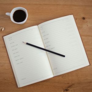 A planner is on a table with a pencil and cup of coffee.