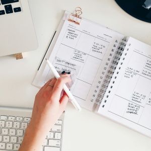 A white woman is at a keyboard with a planner in her white desk and a white ink pen in her hands as she reads the inputs.
