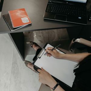 A woman is about to start writing on a paper that's on a clipboard. She is sitting at a desk with 2 books on it.
