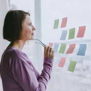 A woman has stick notes taped to a mirror. She is looking at them with her glasses in her hand, up to her chin.