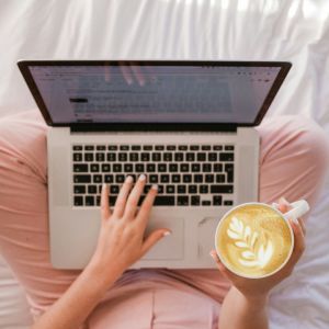 a woman in her bed on a computer, drinking a latte