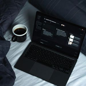 A black laptop and a black cup of black coffee are on a bed with white sheets and a black blanket. The screen on the computer is black and the words are white.