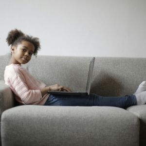 A young girl on a couch with a laptop looking away from it to listen to someone.
