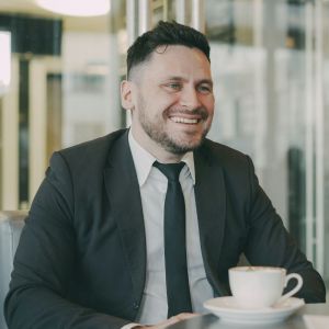 A man in a suit is smiling while sitting at a table with a tea cup in front of him.