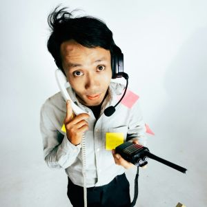 A man with a headset on his head, a phone in one hand, walkie talkie in another, and sticky notes on his shirt.