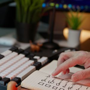 A man with a black and white cover over his keyboard is looking at a to-do list that's written on graph paper.