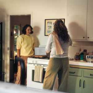 Two women in a home,, standing over a computer; talking while drinking.